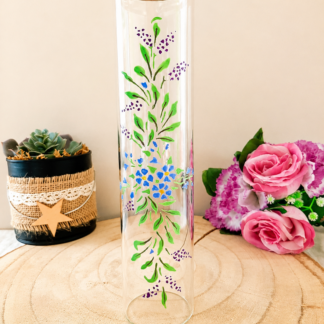Porte-encens cylindrique en verre peint à la main avec bouquet de myosotis bleus et feuillage vert, cerclage doré, posé sur une table en bois avec des roses roses