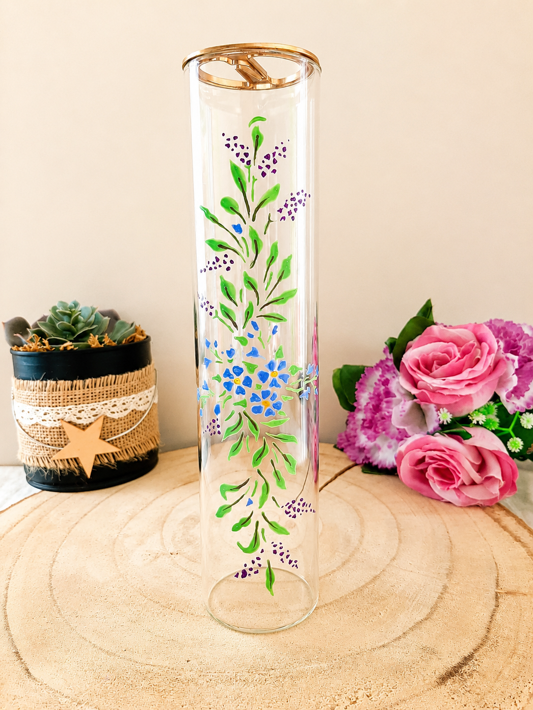 Porte-encens cylindrique en verre peint à la main avec bouquet de myosotis bleus et feuillage vert, cerclage doré, posé sur une table en bois avec des roses roses