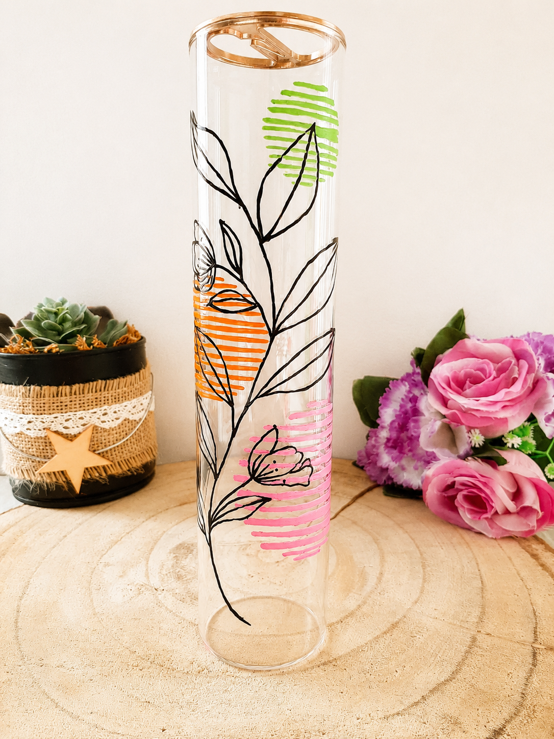 Porte-encens cylindrique en verre peint à la main avec fleurs linéaires noires et touches de couleur vert orange rose, cerclage doré, posé sur une table en bois avec des roses roses