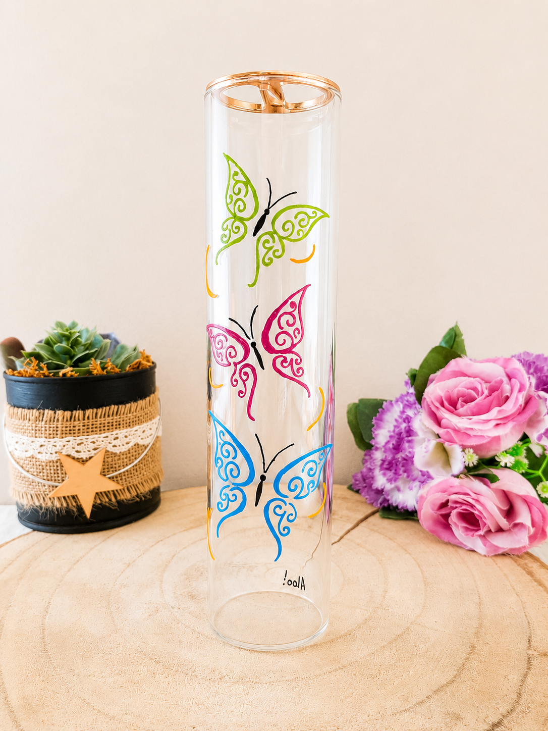 Porte-encens cylindrique en verre peint à la main avec trois papillons colorés vert rose et bleu, cerclage doré, posé sur une table en bois avec des roses roses