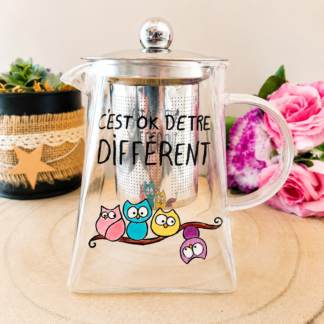 Théière en verre peinte à la main avec inscription C'est OK d'être différent et hiboux colorés sur une branche, filtre inox, posée sur une table en bois avec des roses roses