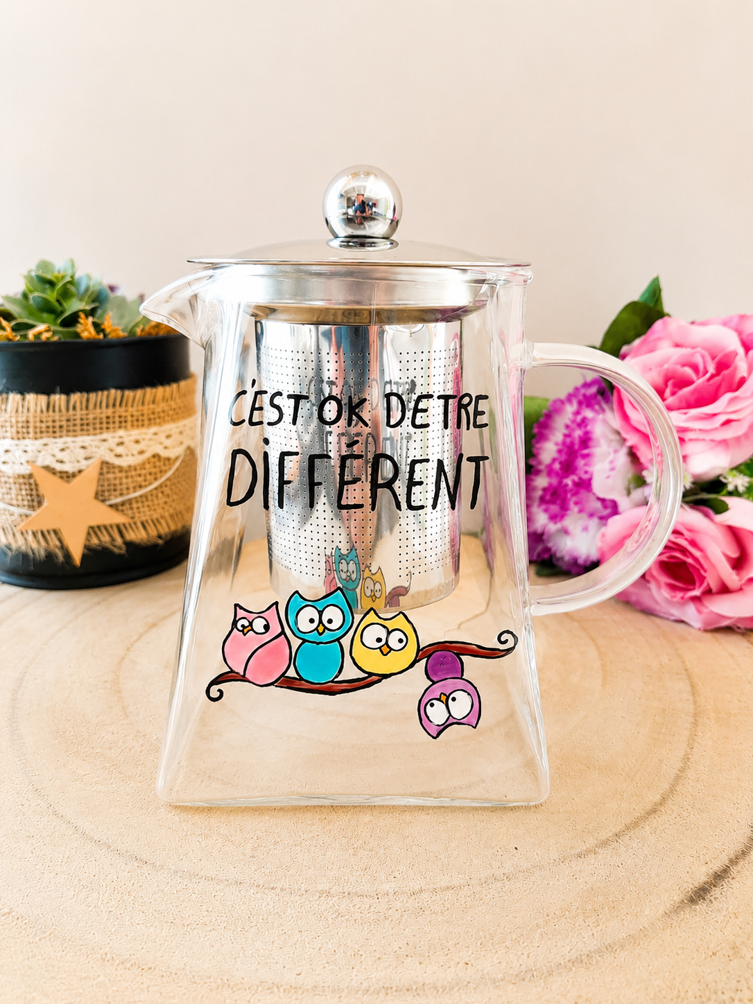 Théière en verre peinte à la main avec inscription C'est OK d'être différent et hiboux colorés sur une branche, filtre inox, posée sur une table en bois avec des roses roses