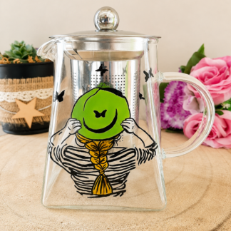 Théière en verre peinte à la main avec femme de dos chapeau vert tresse dorée et papillons noirs, filtre inox, posée sur une table en bois avec des roses roses