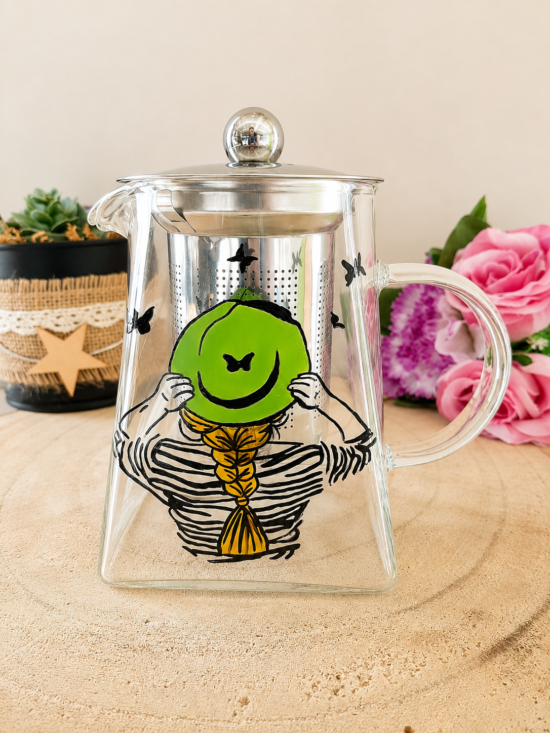 Théière en verre peinte à la main avec femme de dos chapeau vert tresse dorée et papillons noirs, filtre inox, posée sur une table en bois avec des roses roses