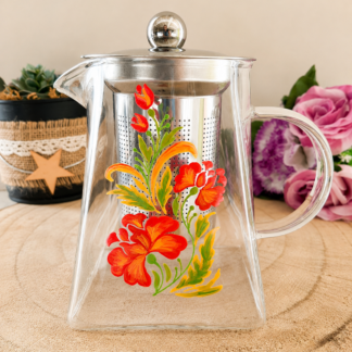 Théière en verre peinte à la main avec bouquet de fleurs rouges et orangées style folklorique, filtre inox, posée sur une table en bois avec des roses roses