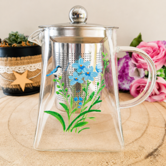 Théière en verre peinte à la main avec grandes fleurs bleues et petit oiseau sur la tige, feuillage vert, filtre inox, posée sur une table en bois avec des roses roses