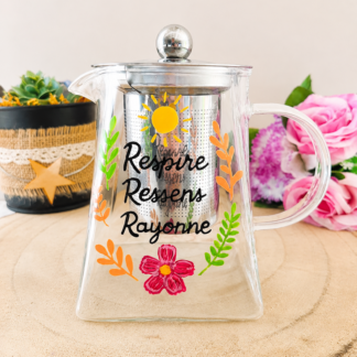 Théière en verre peinte à la main avec inscription Respire Ressens Rayonne et soleil jaune feuillage coloré, filtre inox, posée sur une table en bois avec des roses roses
