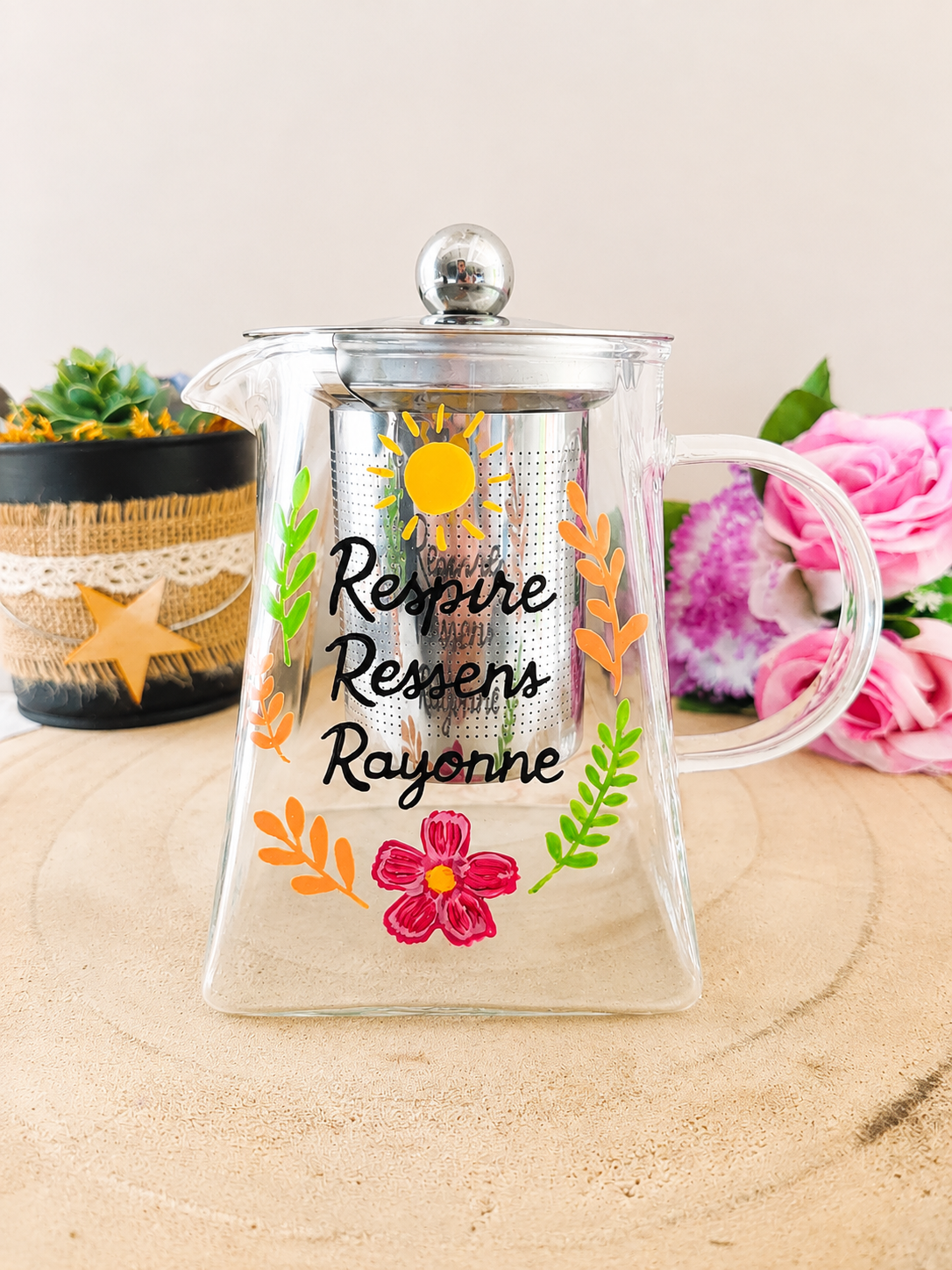 Théière en verre peinte à la main avec inscription Respire Ressens Rayonne et soleil jaune feuillage coloré, filtre inox, posée sur une table en bois avec des roses roses