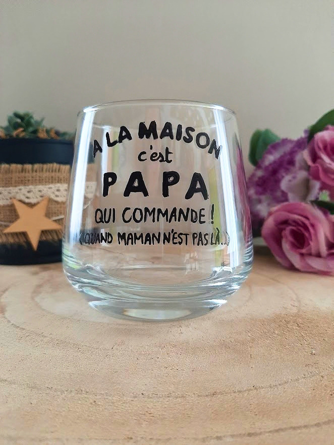 Verre à message humoristique peint à la main "À la maison c'est papa qui commande, quand maman n'est pas là", posé sur une table en bois avec des fleurs roses