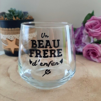 Verre à message peint à la main "Un beau-frère d'enfer" avec petites cornes illustrées, posé sur une table en bois avec des fleurs roses