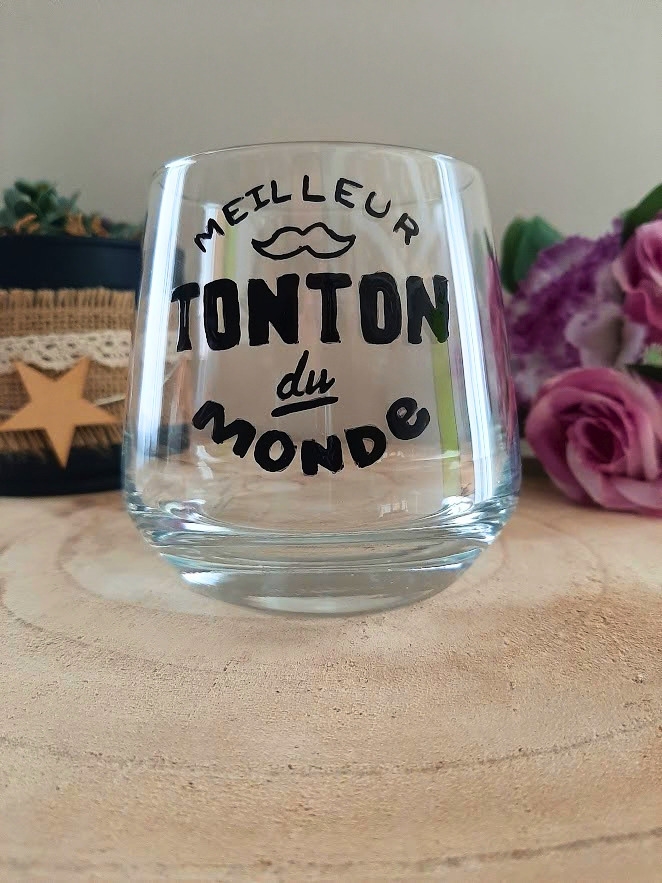 Verre à message peint à la main "Meilleur tonton du monde" avec petite moustache illustrée, posé sur une table en bois avec des fleurs roses