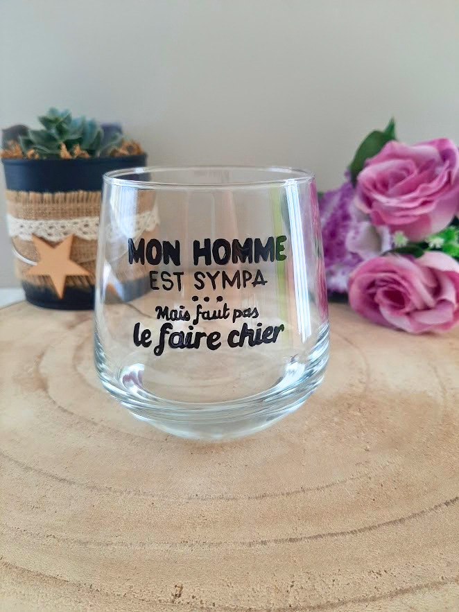 Verre à message humoristique peint à la main "Mon homme est sympa mais faut pas le faire chier", posé sur une table en bois avec des fleurs roses