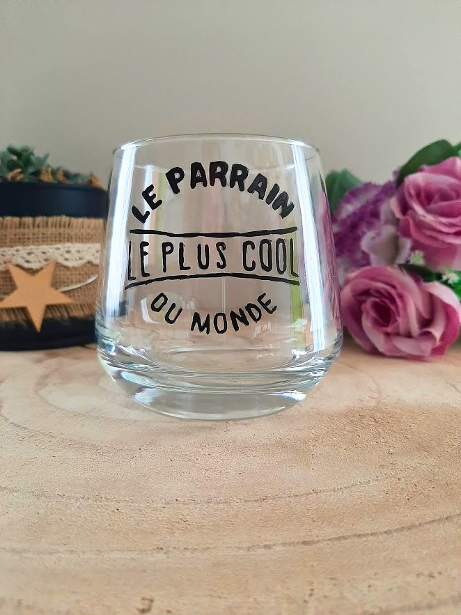 Verre à message peint à la main "Le parrain le plus cool du monde", posé sur une table en bois avec des fleurs roses